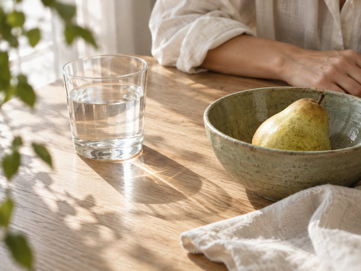 A glass of water, a pear in a sage-green ceramic bowl, and linen on a sunlit oak table